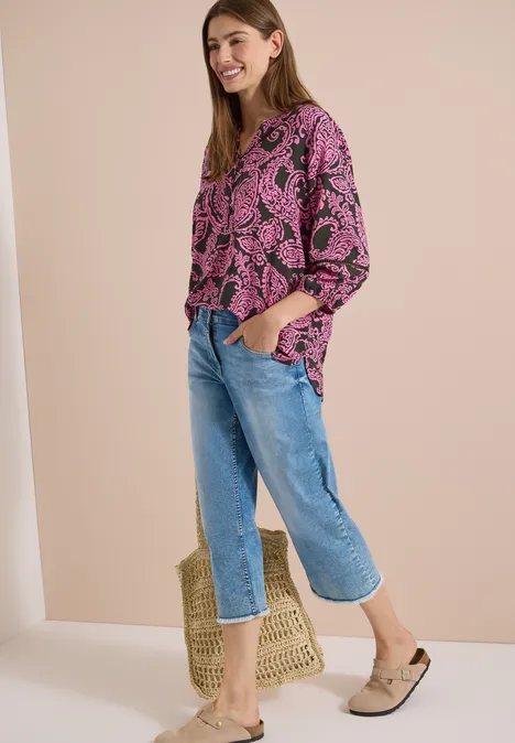 Une femme porte un haut rose imprimé feuilles, jean bleu, mules beiges et un panier en paille.