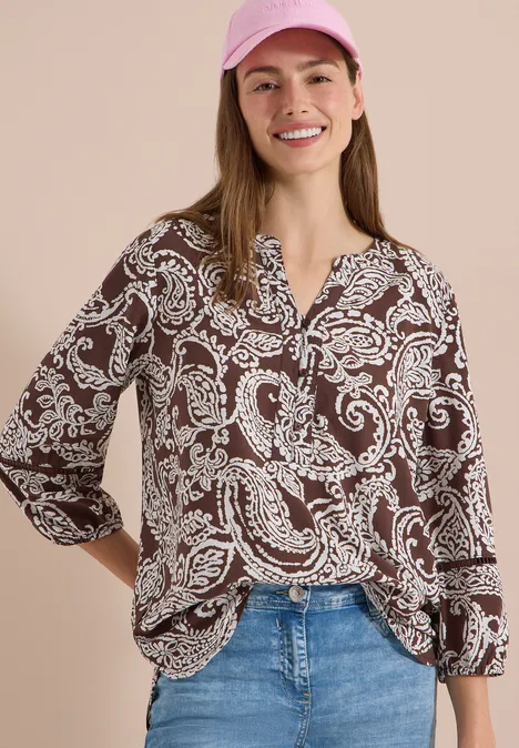 Femme vêtue d’un chemisier marron à motif cachemire blanc, jean et casquette rose sur fond clair.