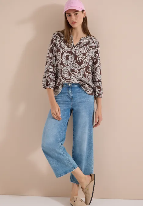 Femme en chemise à motif paisley, jean large bleu et sandales en jute claires devant un mur uni.