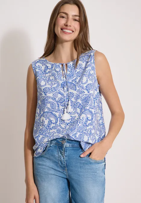 Haut chemisier à col fendu et imprimé vanilla white Femme portant un top sans manches bleu à motif Paisley et jeans, sur fond neutre.
