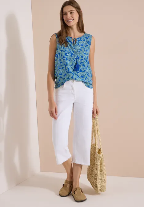 Haut chemisier à col fendu et imprimé surf blue Femme en blouse bleue à motifs, pantalon blanc 7/8, sac en paille et mocassins devant un mur beige.