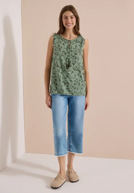 Une femme se tient devant un fond neutre, vêtue d’un top sans manches vert, d’un jean court bleu et de chaussures beiges.