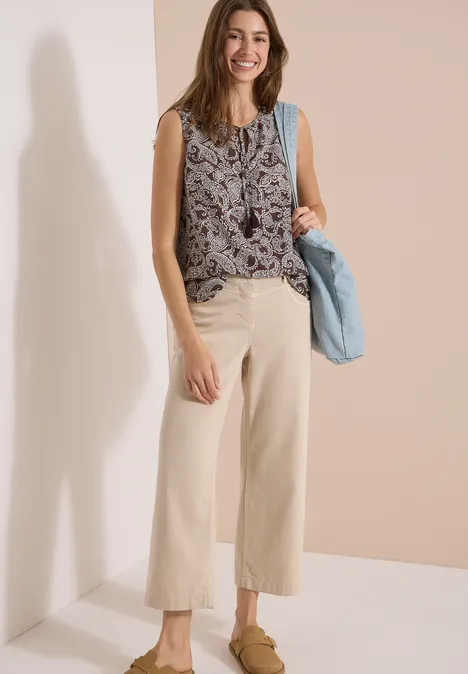 Frau in ärmellosem Top mit Muster, weiten hellen Hosen und blauer Handtasche.