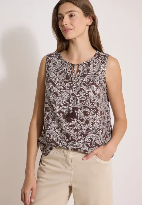 Frau trägt ärmelloses gemustertes Top mit Paisley-Print und Quasten, kombiniert mit beigen Hosen.