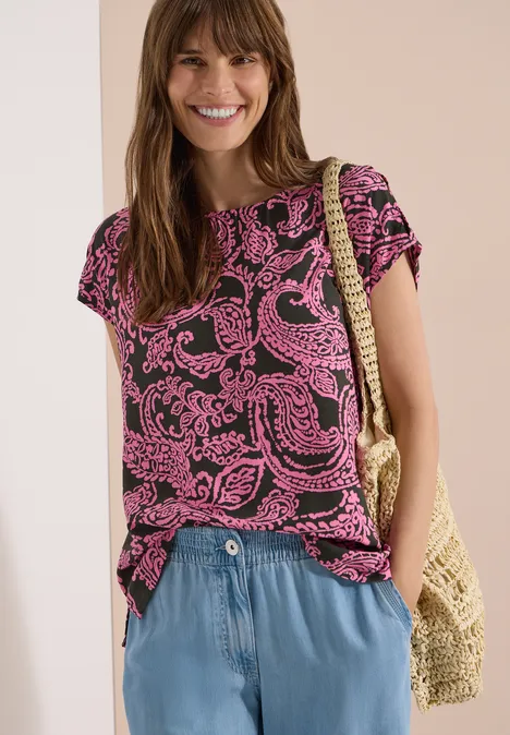 Frau trägt schwarzes T-Shirt mit pinkem Paisley-Muster, blaue Hose und Korbtasche.