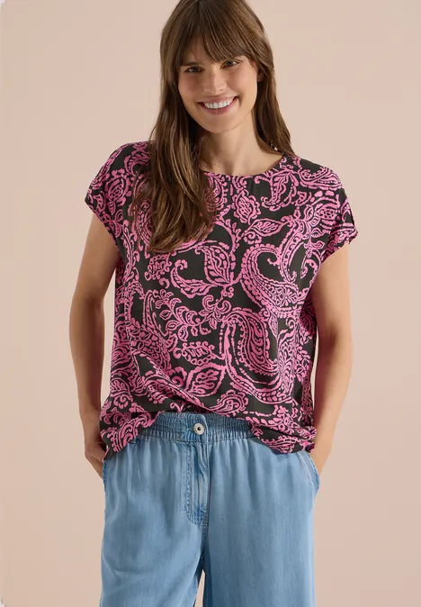 Frau trägt pink gemustertes T-Shirt und hellblaue Jeanshose, steht mit Händen in den Taschen.