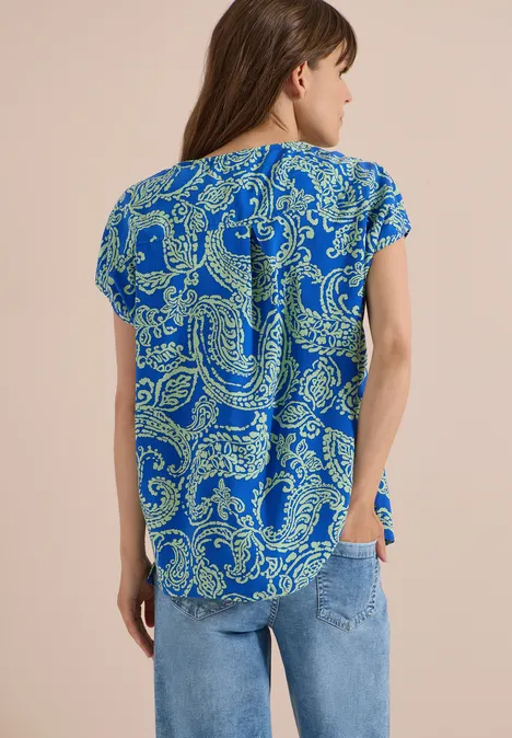 Frau von hinten in blauer Kurzarmbluse mit gelbgrünem Paisley-Muster und Jeans.