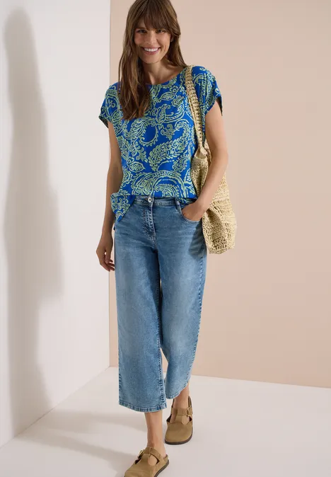 Frau trägt blau gemustertes T-Shirt, helle Culotte-Jeans, Strohtasche und beige Keilsandalen.