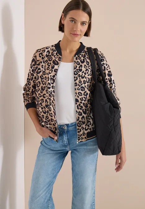 Femme portant une veste bomber léopard, t-shirt blanc, jean et grand sac matelassé noir sur fond clair.