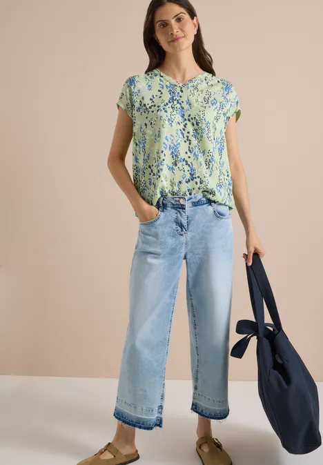 Eine Frau trägt eine geblümte Bluse, weite helle Jeans und hält eine dunkelblaue Tasche.