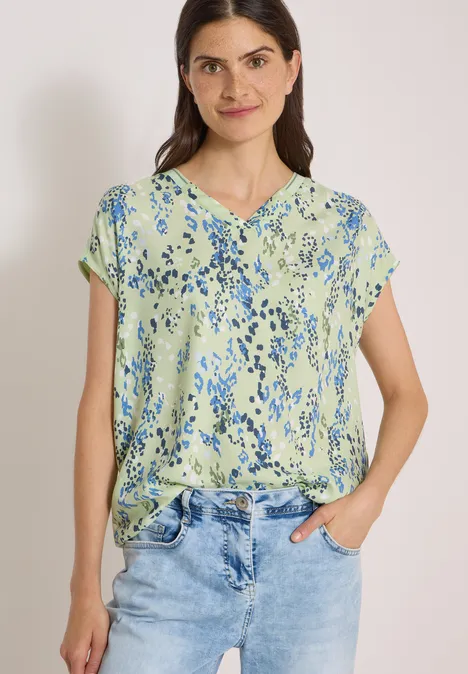 Frau in hellgrüner Bluse mit blauem Blumenmuster und Jeans, lächelnd, Hände in den Taschen.