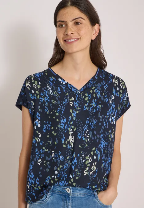 Frau trägt schwarze Bluse mit blauem Blumenmuster, V-Ausschnitt, kurze Ärmel, lächelt, Hände in Taschen.
