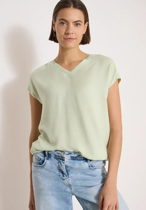 Kurzarm Blusenshirt mit Strukturmix raw matcha lime Frau in locker sitzender, hellgrüner V-Neck-Bluse und hellblauen Jeans vor weißem Hintergrund.