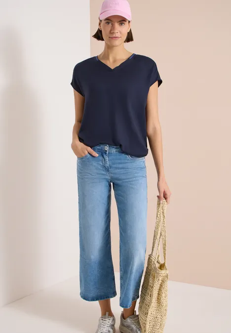 Kurzarm Blusenshirt mit Strukturmix urban dark blue Frau in dunkelblauem V-Shirt und weit geschnittener blauer Jeans mit Strohtasche vor schlichtem Hintergrund.