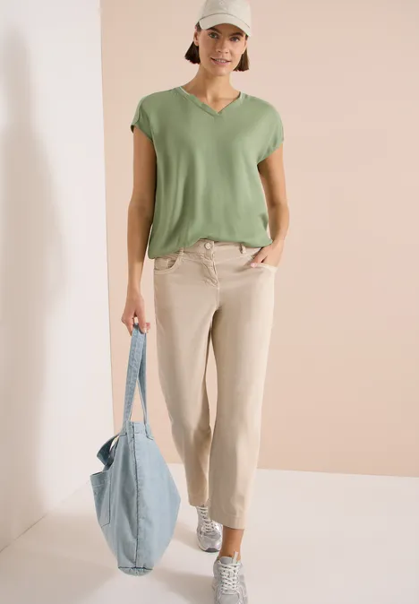 Kurzarm Blusenshirt mit Strukturmix deep matcha green Eine Frau trägt ein grünes T-Shirt, helle Hose, weiße Sneaker und hält eine blaue Tragetasche vor schlichtem Hintergrund.