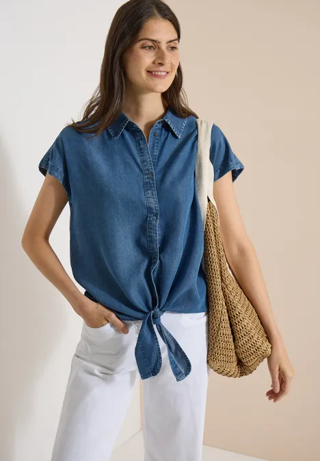 Chemisier à épaules tombantes, effet denim mid blue wash Frau trägt sommerliche blaue Jeansbluse, vorne geknotet, weiße Hose und Korbtasche.