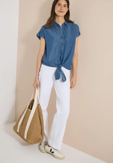 Chemisier à épaules tombantes, effet denim mid blue wash Frau in blauem Kurzarmhemd mit Knoten, weißen Hosen und Strohtasche vor beigem Hintergrund.