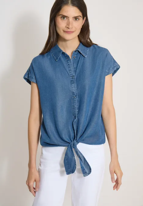 Chemisier à épaules tombantes, effet denim mid blue wash Ärmellose Jeansbluse mit vorne gebundenem Saum und weiße Hose vor einfachem Hintergrund.