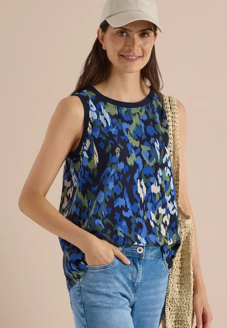 Frau in blau-schwarzem Top, Jeans und Strohtasche, steht lässig mit einer Hand in der Hosentasche.
