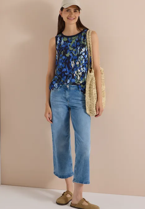 Dame in blau gemusterter äRmelloser Bluse, hellblauen Cropped-Jeans, Strohtasche und Sandalen vor neutralem Hintergrund.