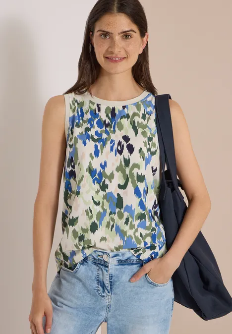 Frau trägt ärmelloses blau-grünes Blumentop, Jeans und hält eine Jacke über der Schulter.