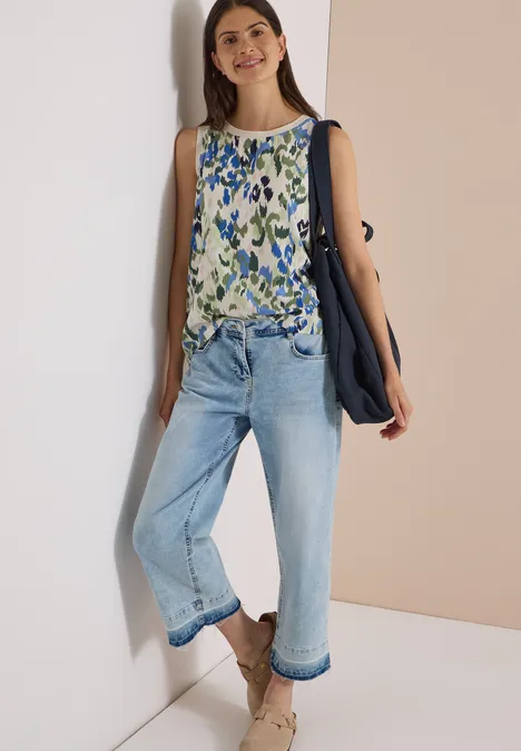 Frau lehnt an Wand, trägt florales Top, weite hellblaue Jeans, braune Sandalen und navyblaue Tasche.
