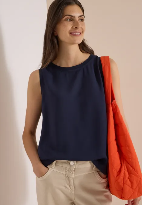 Blusentop mit Rundhals und Rippdetails urban dark blue Frau in marineblauem ärmellosem Top und beigen Hosen, lächelnd mit orangener Stepp-Tasche über der Schulter.
