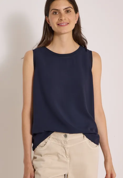Blusentop mit Rundhals und Rippdetails urban dark blue Frau in ärmellosem dunkelblauen Top und beiger Hose lächelt vor neutralem Hintergrund.