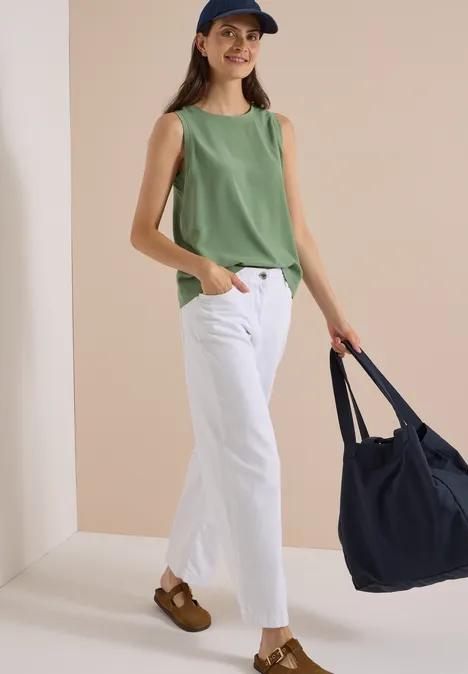 Blusentop mit Rundhals und Rippdetails deep matcha green Frau in grünem Top, weißen Hosen, braunen Clogs und Cap hält navy Tasche.
