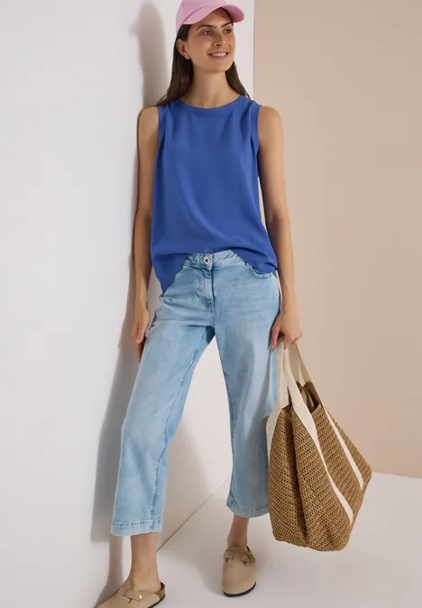 Blusentop mit Rundhals und Rippdetails deep river blue Frau in blauem Top, hellen Culotte-Jeans und rosa Cap mit Strohtasche, die an einer Wand lehnt und lächelt.
