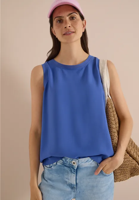 Blusentop mit Rundhals und Rippdetails deep river blue Frau mit pinker Kappe, blauem ärmellosem Top, heller Jeans und Strohtasche vor neutralem Hintergrund.
