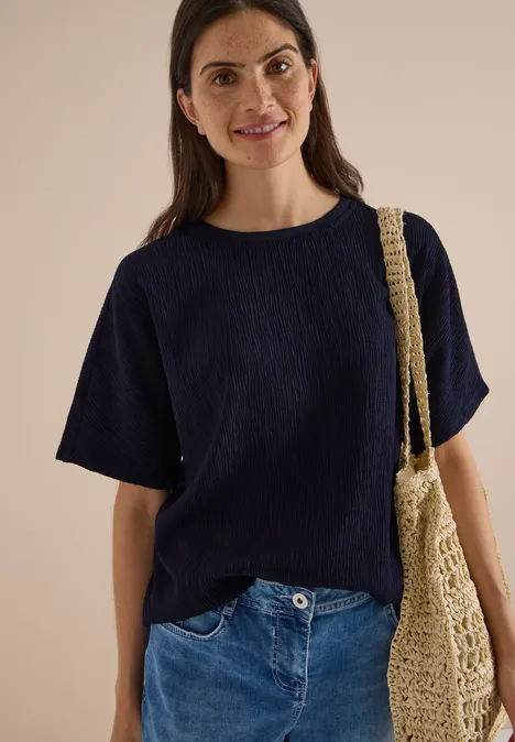 Dropped Shoulder Blusenshirt in Unifarbe urban dark blue Eine lächelnde Frau im dunkelblauen Shirt, Jeans und einer Netztasche vor hellem Hintergrund