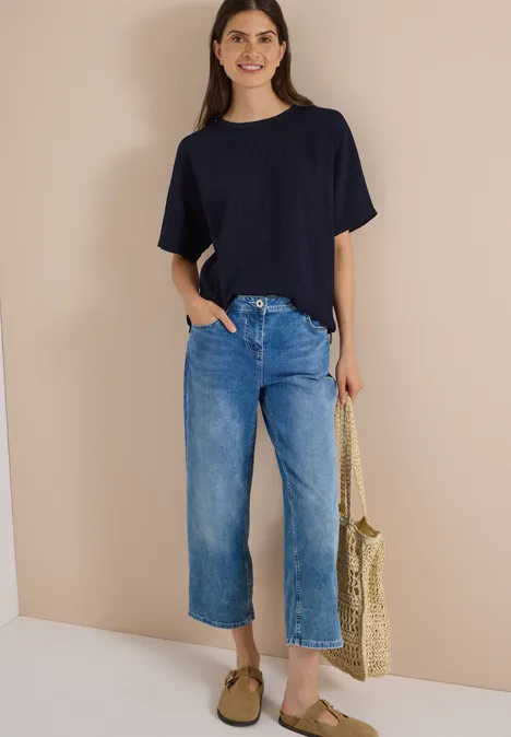 Dropped Shoulder Blusenshirt in Unifarbe urban dark blue Eine Frau trägt ein dunkelblaues T-Shirt, weite blaue Jeans und Sandalen, hält eine geflochtene Stroh-Tasche.
