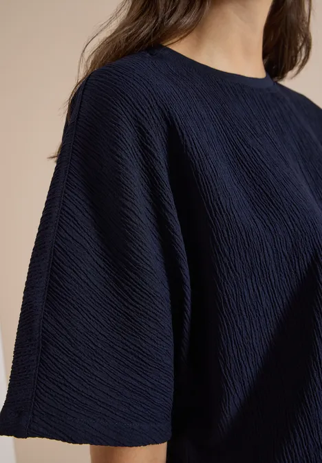 Dropped Shoulder Blusenshirt in Unifarbe urban dark blue Teilaufnahme einer Person in einem dunkelblauen, strukturierten, weiten Kurzarmshirt vor beigem Hintergrund.