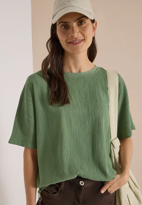 Dropped Shoulder Blusenshirt in Unifarbe deep matcha green Lächelnde Frau in grünem, strukturiertem T-Shirt, beige Cap und Canvas-Tasche vor neutralem Hintergrund.