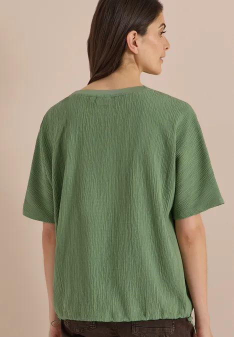 Dropped Shoulder Blusenshirt in Unifarbe deep matcha green Frau von hinten in locker sitzendem, kurzärmeligem, grünem, leicht gekräuseltem Shirt vor beigem Hintergrund.