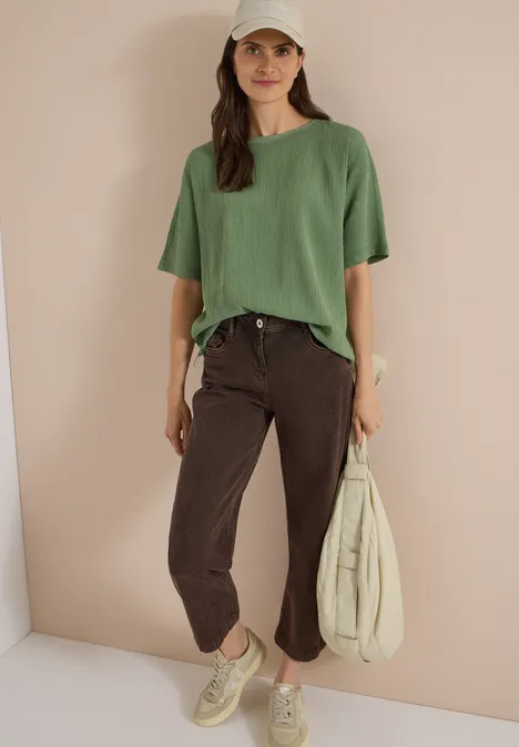 Dropped Shoulder Blusenshirt in Unifarbe deep matcha green Frau in grünem Shirt, braunen Hosen, Sneakern und weißer Cap lehnt vor beiger Wand, trägt Tasche.