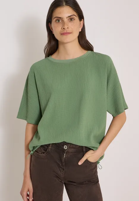 Dropped Shoulder Blusenshirt in Unifarbe deep matcha green Frau in grüner Bluse und dunkler Hose, eine Hand in der Tasche, vor hellem Hintergrund.