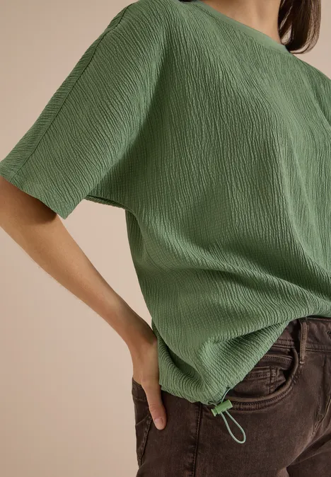 Dropped Shoulder Blusenshirt in Unifarbe deep matcha green Eine Person trägt ein grünes, strukturiertes Kurzarmshirt mit Kordelzugsaum und braune Hosen