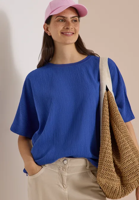 Dropped Shoulder Blusenshirt in Unifarbe deep river blue Frau in blauer Bluse, rosa Hut und beiger Hose, lächelt und trägt eine geflochtene Tasche.