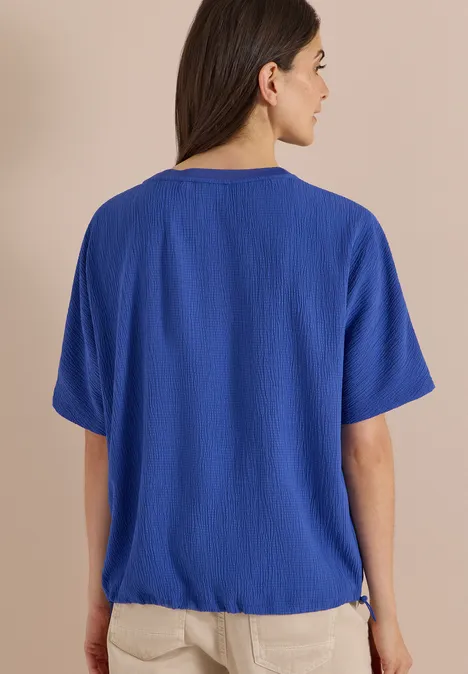 Dropped Shoulder Blusenshirt in Unifarbe deep river blue Eine Frau mit dunklen Haaren trägt eine blaue Bluse und helle Hose, von hinten vor beigem Hintergrund.