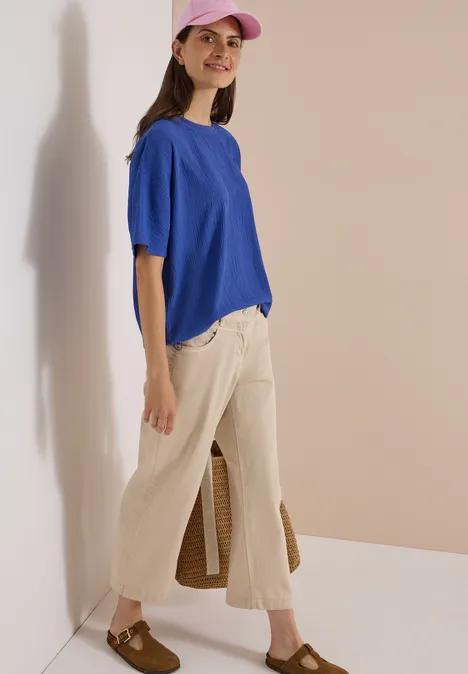 Dropped Shoulder Blusenshirt in Unifarbe deep river blue Eine Frau mit rosa Baseballcap, blauem T-Shirt, beigen Culottes und braunen Loafers vor hellem Hintergrund.