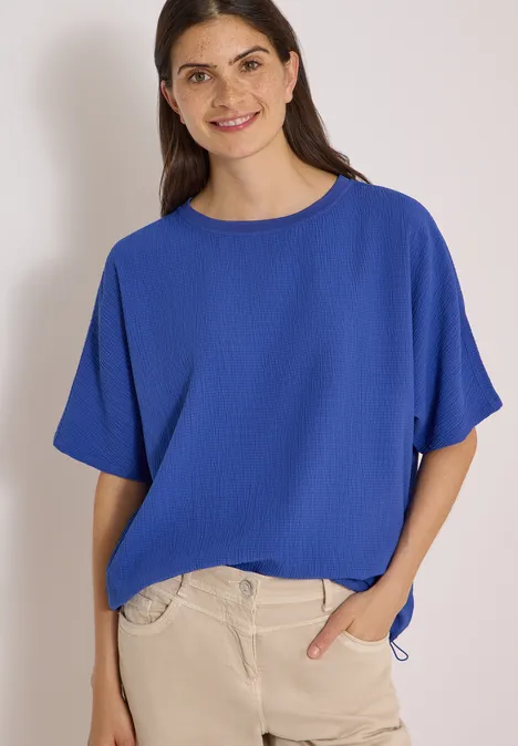 Dropped Shoulder Blusenshirt in Unifarbe deep river blue Frau in lockerem blauen T-Shirt mit kurzen Ärmeln und heller Hose, lächelnd vor hellem Hintergrund.