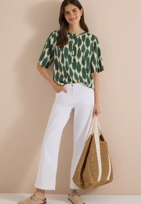 Kurzarm Bluse mit Rundhals und Print vanilla white Frau trägt grün gemustertes Shirt, weiße weite Hose und hält eine große geflochtene Strohtasche.