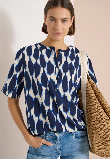 Kurzarm Bluse mit Rundhals und Print urban dark blue Frau in kurzärmeliger blau-weißer Bluse, Jeans und großer geflochtener Einkaufstasche vor neutralem Hintergrund.