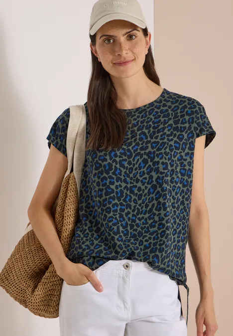 Frau in blau-grünem Leo-Shirt, weißer Hose und Strohtasche, mit Cap, lässig lächelnd