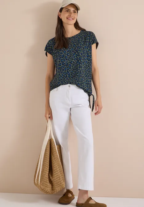 Frau mit gemusterter Bluse, weißer Leinenhose, Strohtasche, Cap und braunen Loafers.