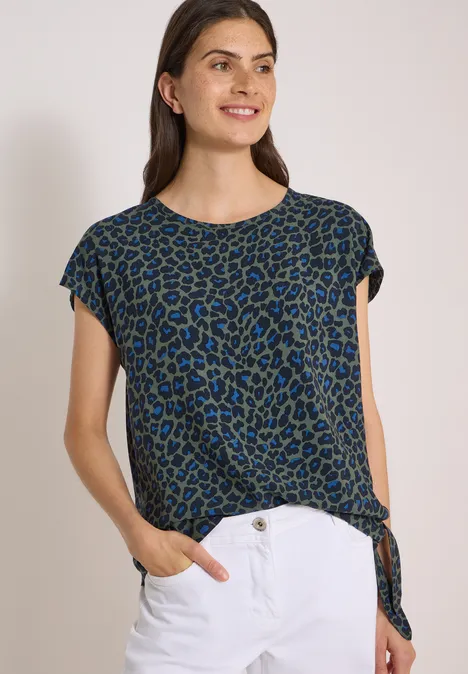Frau trägt blau-grünes Leo-Shirt mit seitlicher Knoten-Optik und weiße Hose, lächelnd.