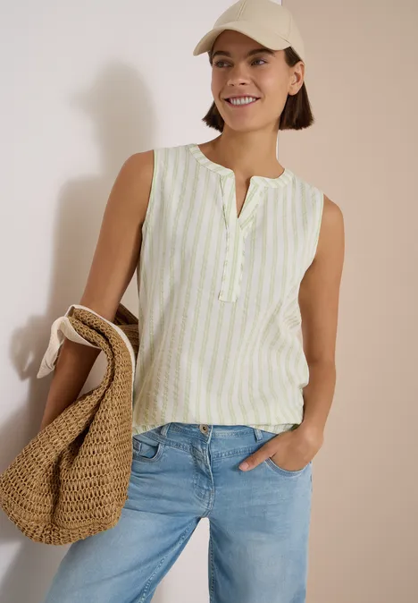 Seersucker Blusentop mit Streifen raw matcha lime Frau in gestreiftem ärmellosem Top, Jeans und Strohtasche, lächelnd vor heller Wand.