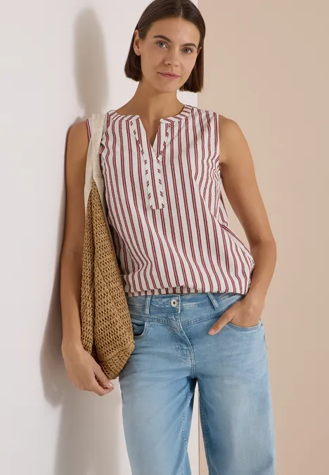 Seersucker Blusentop mit Streifen russet red Frau lehnt lässig an Wand, trägt gestreiftes ärmelloses Top, helle Jeans und Korbtasche.